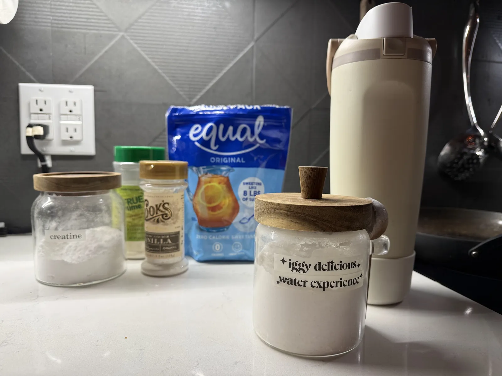 a kitchen counter at night: a jar of creatine, a shaker of True Lime, a jar of vanilla powder, a bag of Equal, a big beige water bottle, and in front a glass jar of white powder labelled ✦iggy delicious✦ ✦water experience✦